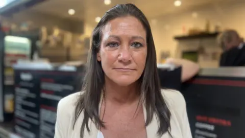 Emma Layton looks directly at the camera. She has long dark hair and blue eyes. She is wearing a cream-coloured jacket. Behind her in soft-focus is a cafe counter.