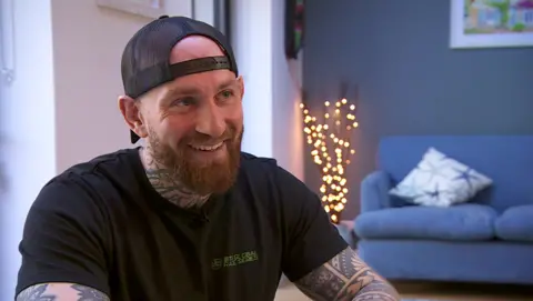 Marc Rhodes is smiling and has a baseball cap on back to front. He is wearing a black T-shirt and has tattoos on his neck and arms. 