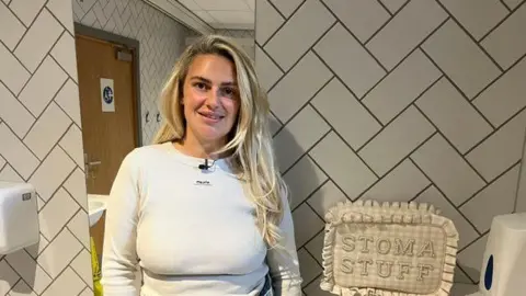 Jenna Robinson is smiling into the camera. She has long blonde hair and is wearing a white top. She is standing in a public toilet which has white tiles on the wall and a long mirror. On a shelf next to her is a beige gingham bag which says "STOMA STUFF" in capital letters. 