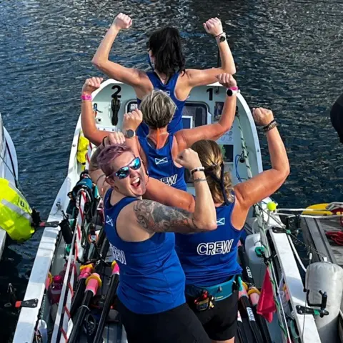 Scotland the Wave The women are standing in their boat and striking a pose by raising their arms and showing their muscles.