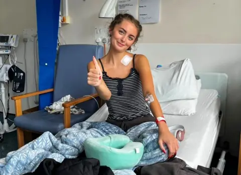 Erin Lavery Person sitting upright in a hospital bed with a blanket over their legs, giving a thumbs-up gesture. Medical monitoring equipment, a chair, and hospital room furnishings are visible in the background. A travel neck pillow rests on the bed in the foreground.