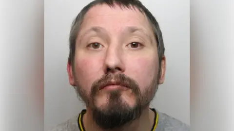 A police mugshot of a man with short black hair, a black beard and a grey top.