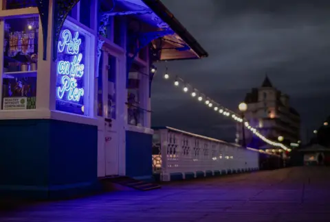 Llandudno pier at night. Photo taken from the pier with a Pets on the Pier Shop  sign lit up in a blue shop window and the railings lit by overhead string lights.