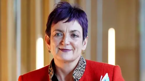 PA Media Angela Constance has short purple hair and smiles while looking off to the side. She wears a red jacket with a leopard print collar