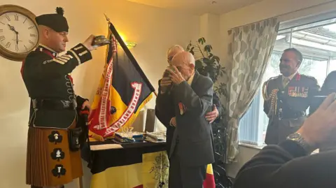Mr Irwin wearing a suit with his military medals stands up in front of a table which has cake and whiskey sitting on it. He is surrounded by military representatives, and another man has his arms around him. The flag on the table and hanging up is yellow and black, and the military representatives are wearing official uniform.