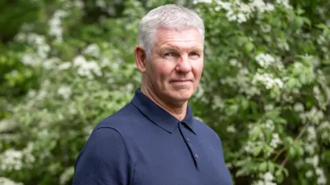 A man with grey hair and a dark blue t-shirt looks at the camera 