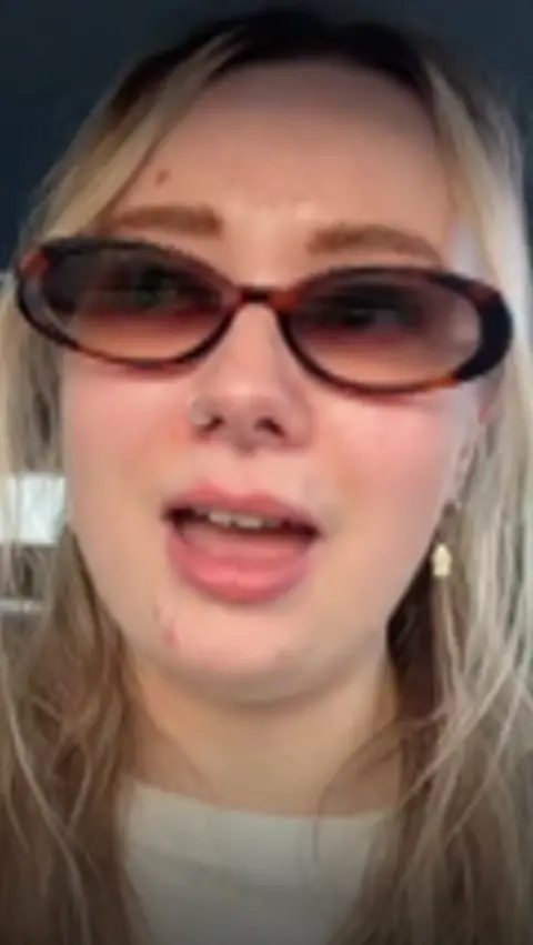 A close-up image of a woman with blonde hair and sunglasses videoing herself talking