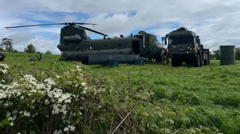 BBC/Emma Davies A closer shot of the aircraft in the field from the above picture. It is dark green and the front rotor blade has been removed. A military truck is parked in front of it.