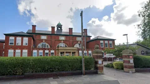 Google Clifton Community School exterior - a red brick building with bushes on its perimeter and a blue but cloudy sky above.