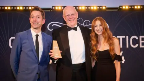 NHS Sussex An elderly, bald man in a black tuxedo and glasses holding a wooden award and smiling at the camera with a younger man in a navy suit and a younger, ginger woman standing next to him.