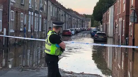 A policeman looks away from the camera, down a street of terraced houses which has been flooded.
