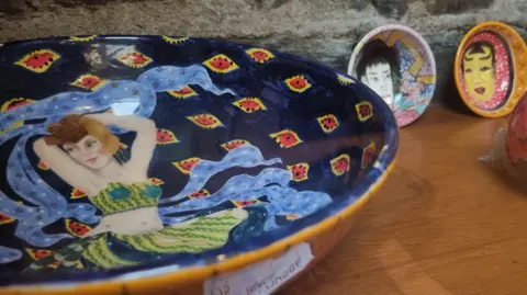 SALLY BLACK A blue red and yellow ceramic bowl with a woman wearing a green dancing outfit of a crop top and skirt. She has brown hair and her hands are behind her head with thick blue ribbons flowing around her. There are two smaller bowls with faces painted on them to the right on a wooden table.