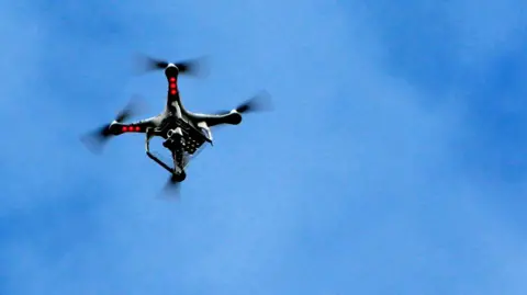 A drone flying in a blue sky. It is black, has four propellers and red lights on it.