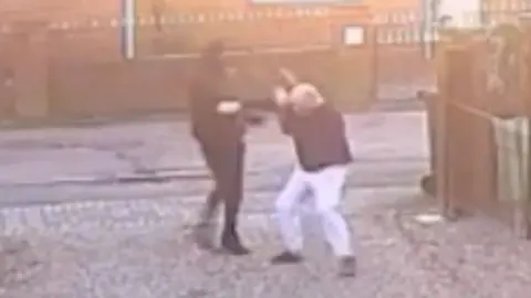 A person dressed in all black hitting a man, wearing white trousers and a dark top, with a wooden plank on the driveway of a house.