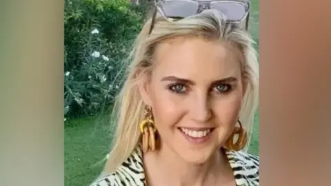 A smiling woman with long blonde hair, hoop earrings, and sunglasses resting on her head stands outdoors in front of green grass and flowering bushes. She wears a patterned outfit and look directly at the camera.
