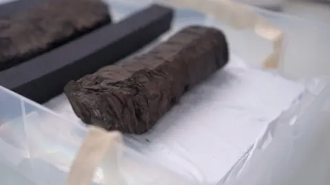 Bodleian Library A scroll from ancient city of Herculaneum sitting in a plastic tray on a desk at the Bodleian library. It is about about 15cm long and about 3cm wide. It is dark brown and appears very charred, resembling a lump of charcoal.