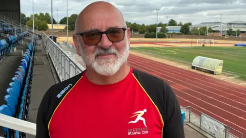 Phil Garner - a bald man with a white moustache, stubble and beard - is wearing sunglasses and smiling at the camera. He is wearing a red and black Grantham Athletics Club top. Behind him is a red running track and blue plastic seats which are empty.