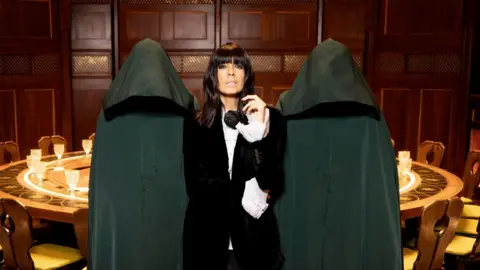 Claudia Winkleman is pictured next to two people wearing black cloaks in front of theround table which is used in the TV show the Traitors