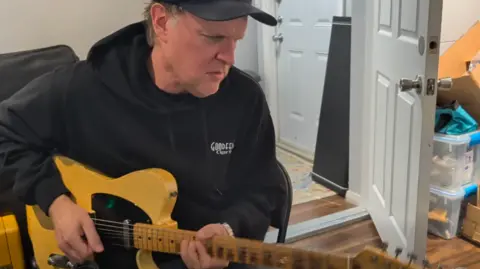 Fiona Long Guitarist Joe Bonamassa sat in a small room playing a yellow and black Fender Telecaster guitar. He is wearing a black hoodie and black cap.