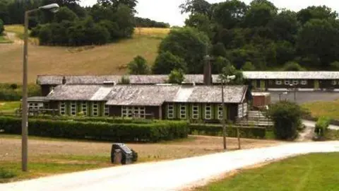 Rows of dormitary style buildings with slate rooves and green window frames sit in a green space in front of a gentle slope lined with trees.