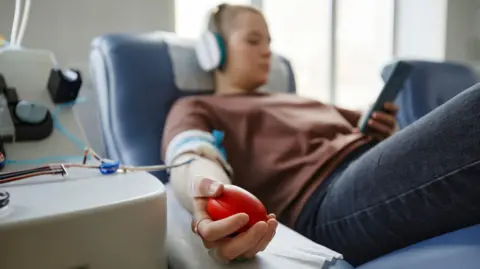 A stock photo of a woman sat in a blood donation chair. She's wearing dark blue jeans and a brown jumper, and has a pair of headphones in and a mobile phone in her left hand. Just her right hand is in focus - where she's squeezing a stress ball to keep up her blood pressure. Her right arm has plastic tubes coming out of it.
