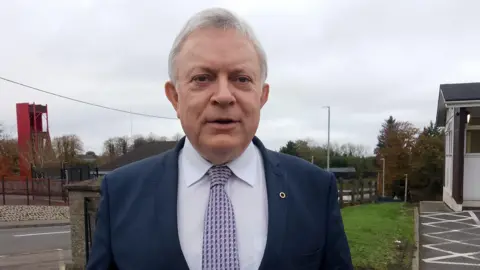 Man standing outside. He is wearing a suit and looking at the camera. Behind him a road and some grass and a fence.