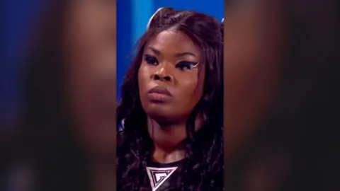 A close-up image of woman with black hair wearing a Gladiators top. She is looking off-screen and has black eye make-up on.