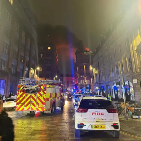 Carla Jenkins A stone building, with smoke rising above it. There are emergency service vehicles in the foreground with brightly coloured lights flashing.