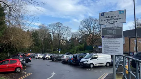 Photograph of the car park at Bramhall Precinct. There are dozens of cars in the picture and a sign explaining the tariffs.