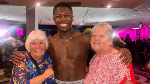 DeReiss Gordon/ Man Like DeReiss Professional wrestler Man Like DeReiss poses in the middle with his arms around Susie and Lynn. Susie, has short white hair and wears a blue flowery top, DeReiss is topless with a silver chain, and Lynn wears a red and white top. All three are smiling at the camera. 