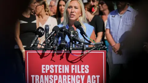Greene standing at podium, speaking into microphone with crowd behind her, and a sign that says "Epstein files transparency act"