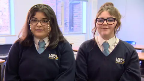 Two teenage girls, both wearing glasses, sitting side by side in a school classroom