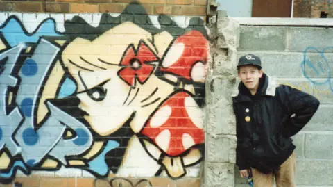 Boy wearing baseball cap and black jacket is standing next to a painted wall with a cartoon depiction