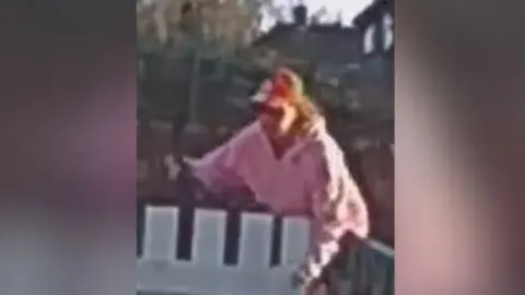 South Yorkshire Police A grainy CCTV image of a woman in a pink hoodie and red baseball cap leaning over a white gate.