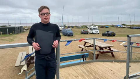 Alex Hawtin is wearing a wetsuit and some grey jogging bottoms over it. He stands at the rail at Snettisham Beach Sailing Club overlooking a line of boats and the sea