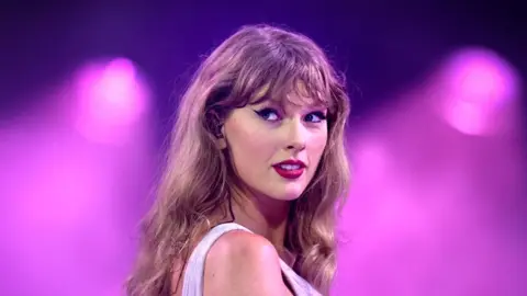 Close up headshot of Taylor Swift performing onstage during The Eras Tour at Wembley Stadium. She has long hair and is looking back over her shoulder.