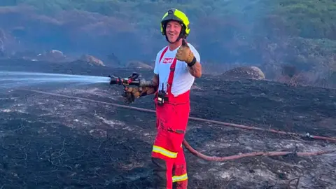 Truro Community Fire Station A firefighter wearing his uniform in the heat while spraying a hose to put out a wildfire on a patch of burnt out ground in Cornwall.