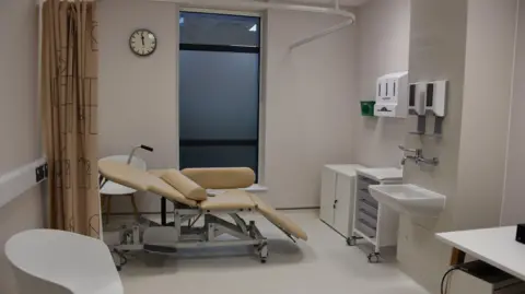 NHS Somerset FT A hospital room, with a bed at the end of the room. There is a curtain hanging down as well as a sink and set of cabinets.