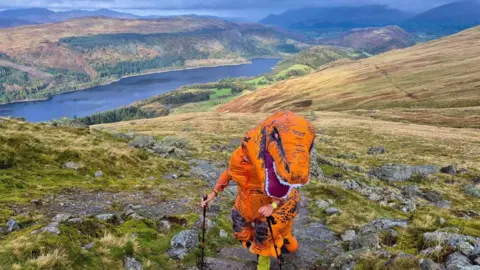 Jay Mistry dressed in an orange t-rex costume, climbs steps in the lake district with two walking poles behind him is a large lake and multiple mountain ridges.