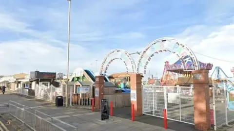Entrance to a small seaside amusement park with white metal gates and brick pillars, decorative archways overhead, and rides visible inside under a bright sky.