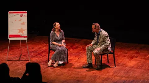 Handout Isobel Jeffery sat in a chair on stage with another person in a chair opposite. Isobel is wearing a long dress and the other person a suit.