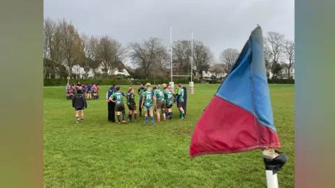 The pink python club versus the emerald pythons. There are players in both pink and green kits in the image. They're stood in two team huddles on the pitch at Hove recreation ground.