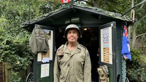BBC A man stands to attention outside a garden shed. He is wearing khaki combat fatigues and hard hat. hanging from the green doors of the shed are a khaki army bag and binoculars. There are posters on it about airplanes and a Flying Tigers bomber jacket. There is foliage in the background.