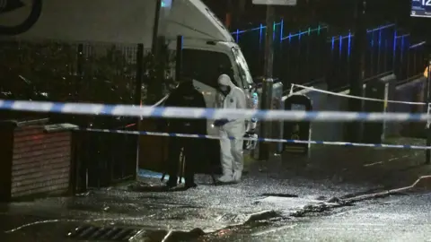 Pacemaker A forensics officer at the scene of a shooting, it is nighttime and the ground is wet, there is a man in a white suit stood beside a police man, who is facing away from the camera in dark clothing. There is police tape across the road.