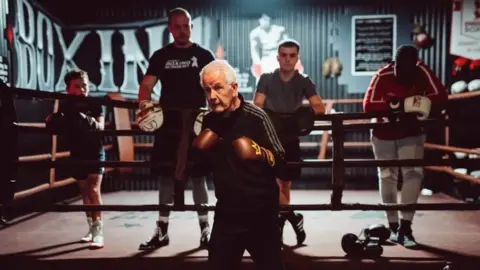 Paul Gilfeather Frank Gilfeather está justo afuera del ring en una postura de boxeo vistiendo un chándal negro con un borde blanco. También lleva guantes de boxeo marrones con coronas doradas y detrás de él hay cuatro estudiantes que están parados dentro del ring apoyados en las cuerdas.