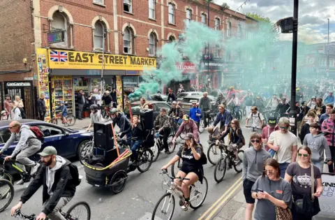 People taking part in the Bristol drum and bass bike ride travel down Bedminster Parade with a man holding a smoke flare sending out green smoke