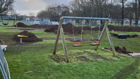 Mark Mather A set of swings is in the foreground of the picture and behind there are large mounds of dug-up earth. Fencing is all round the site.