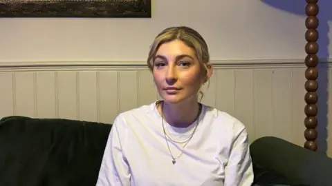 Woman with blonde hair and a white long-sleeved T-shirt sitting on her sofa 