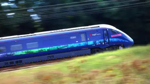 A blue Hull Trains speeding along railway tracks. There is a motion blur in the background and foreground. The train features branding with "Hull Trains" on its side.