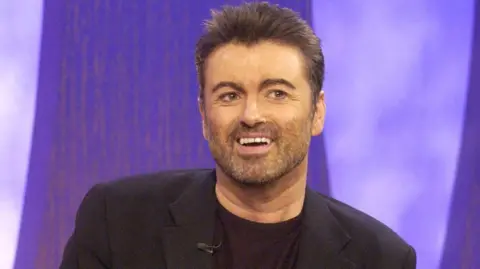 George Michael on the Parkinson chat show in 2004. He is wearing a black jacket and is smiling.